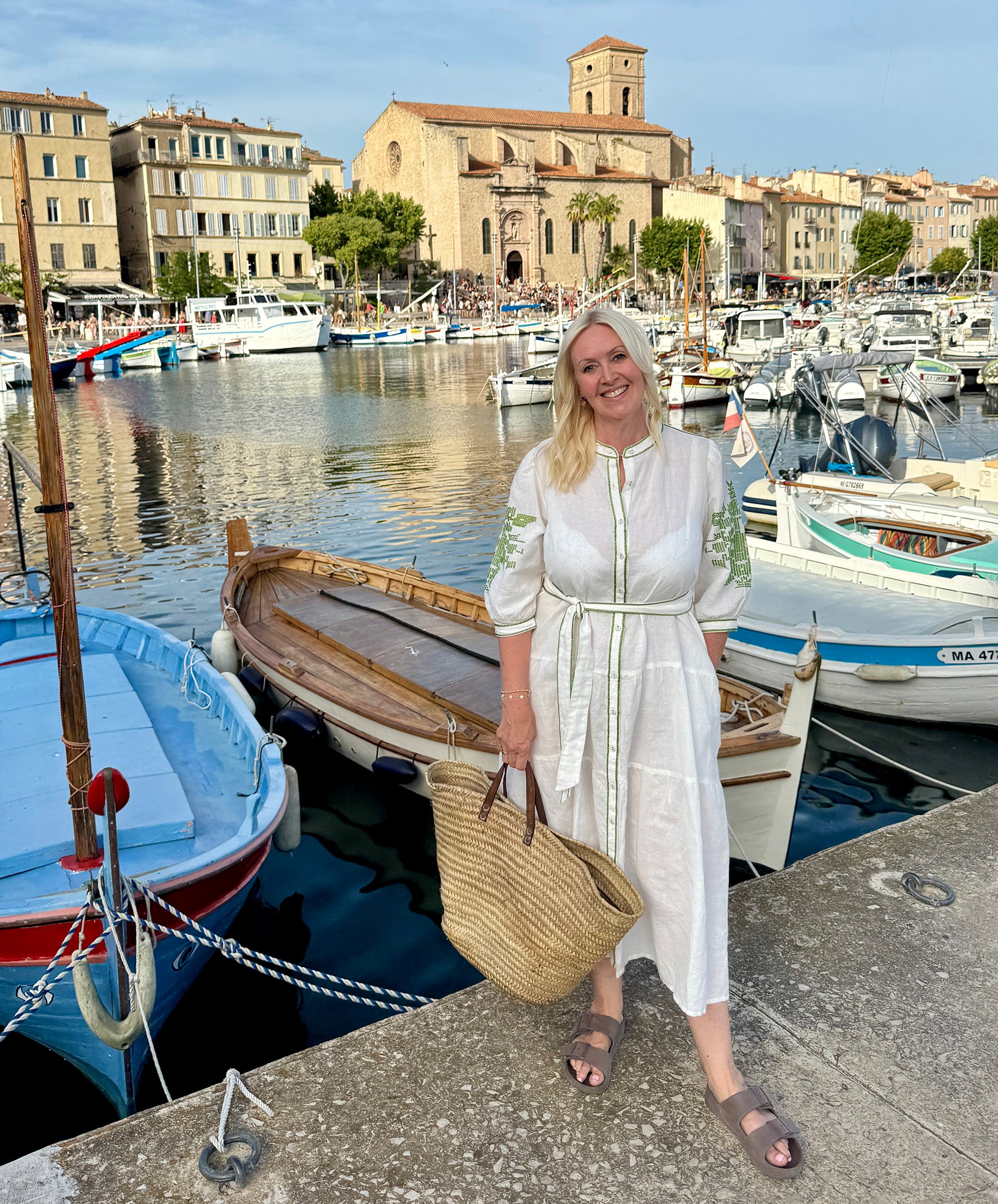 Influencer  @shutters_and_stone wearing the Pine white dress with olive green embroidery, stood next to an old marina.