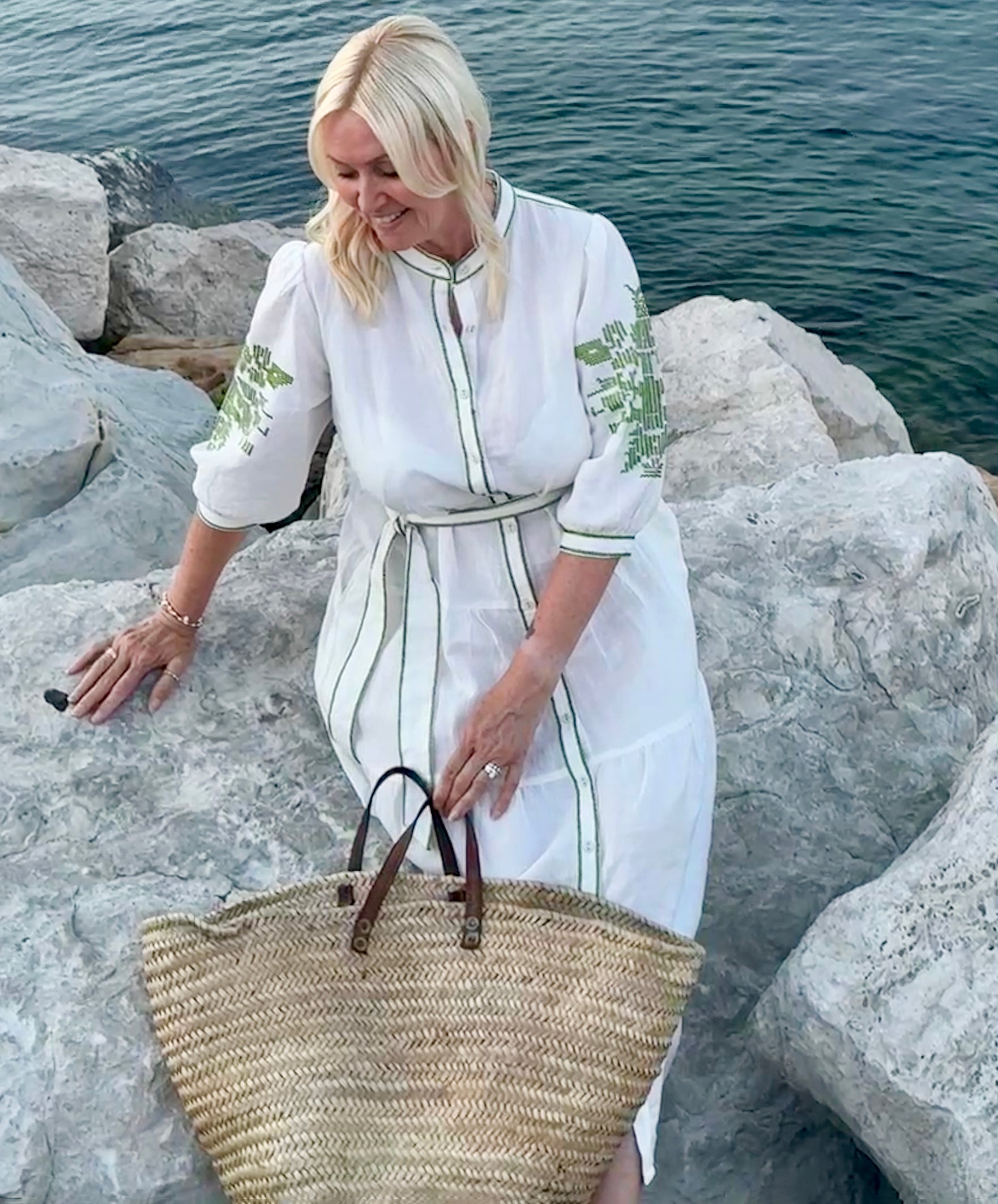 Influencer @shutters_and_stone wearing the Pine white dress with olive green embroidery, sitting on a rock by the sea with a straw bag in hand.