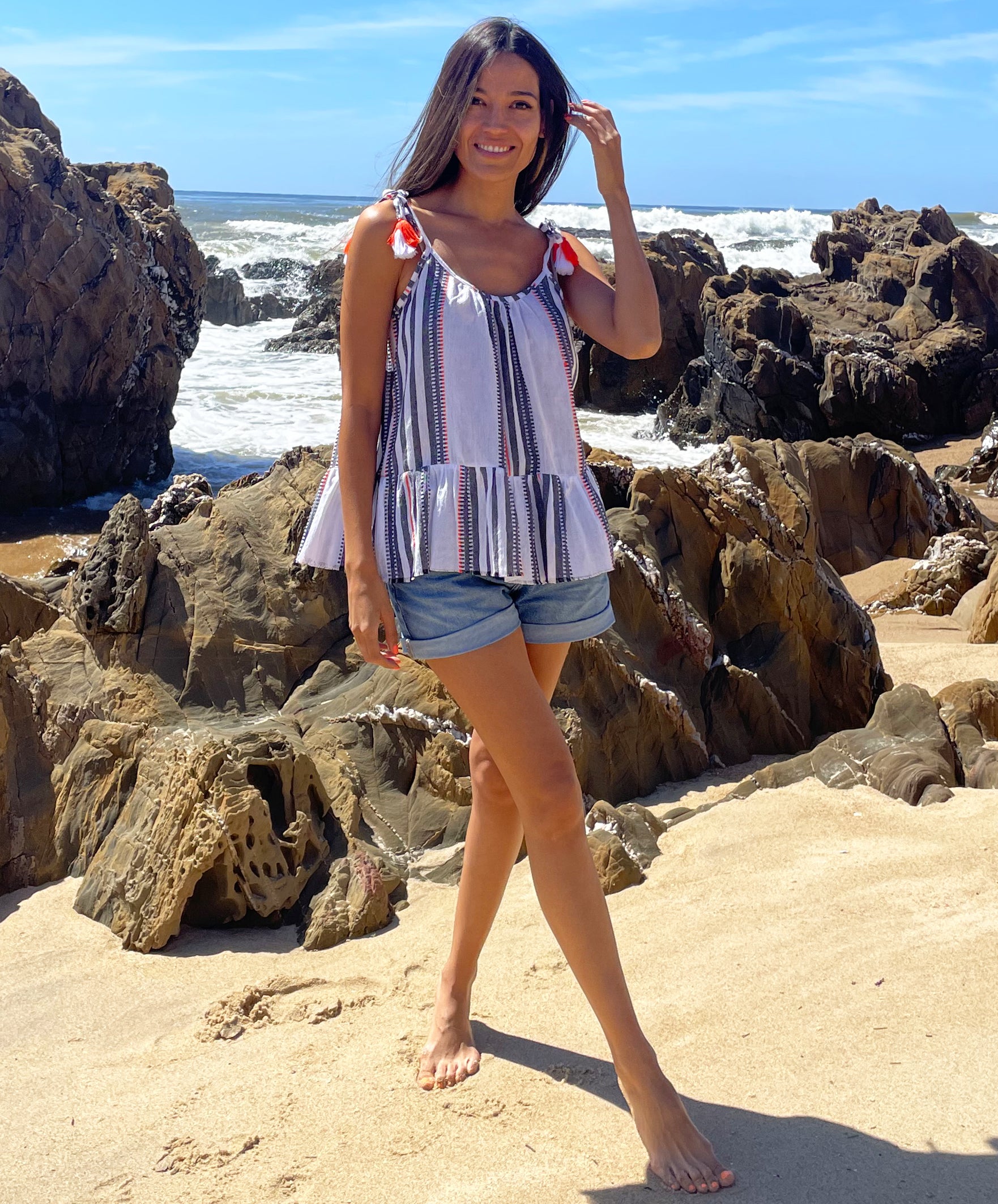 A  model stood on a beach wearing a Rosebud by Rose and Rose Bergerac Top and denim cut off shorts. 