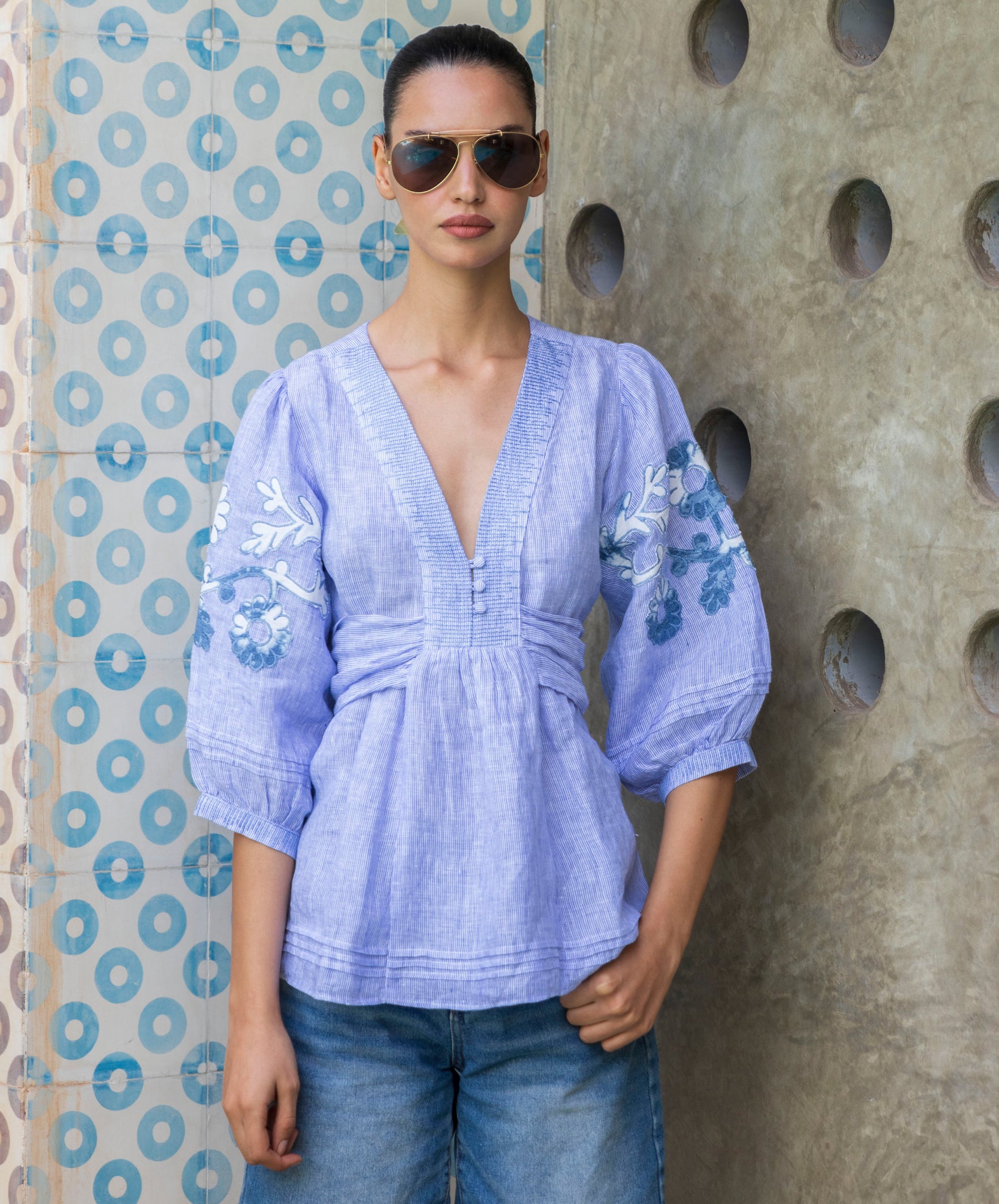 Woman wearing a blue striped Blossom  embroidered blouse and sunglasses against a textured wall.