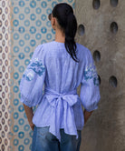 Woman wearing a blue striped Blossom embroidered blouse against a patterned wall.