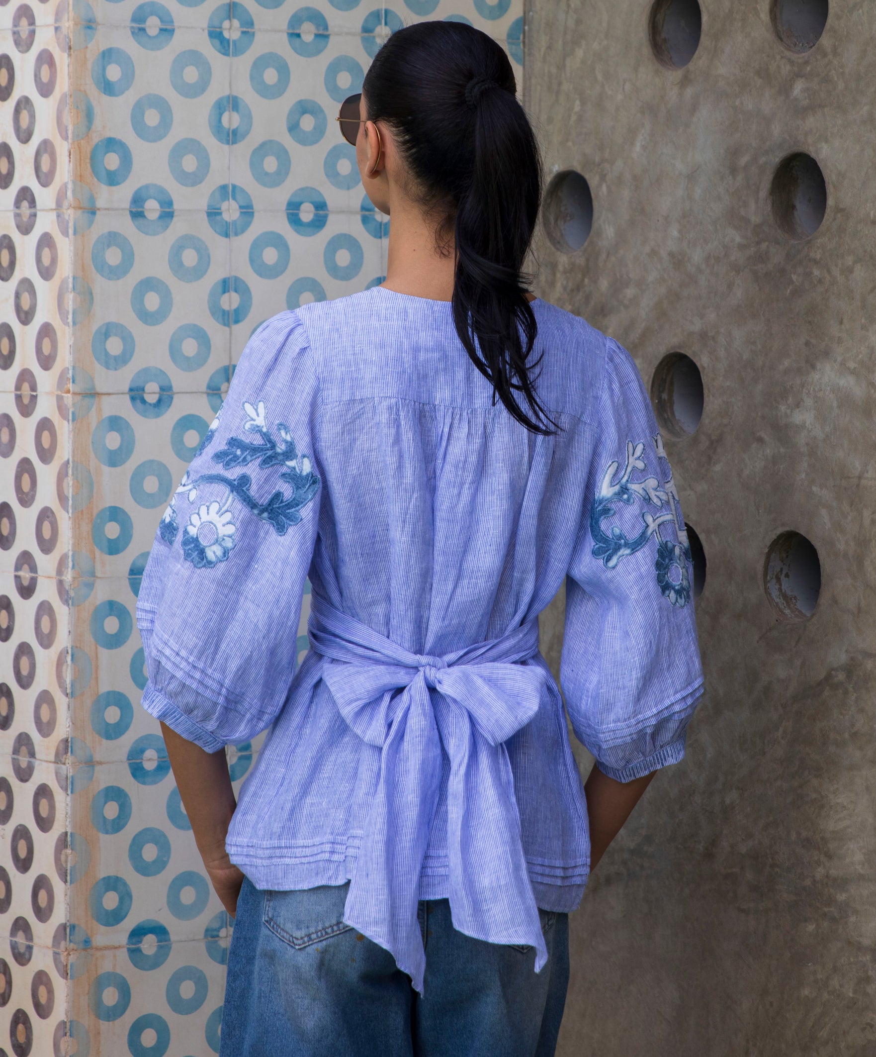 Woman wearing a blue striped Blossom embroidered blouse against a patterned wall.