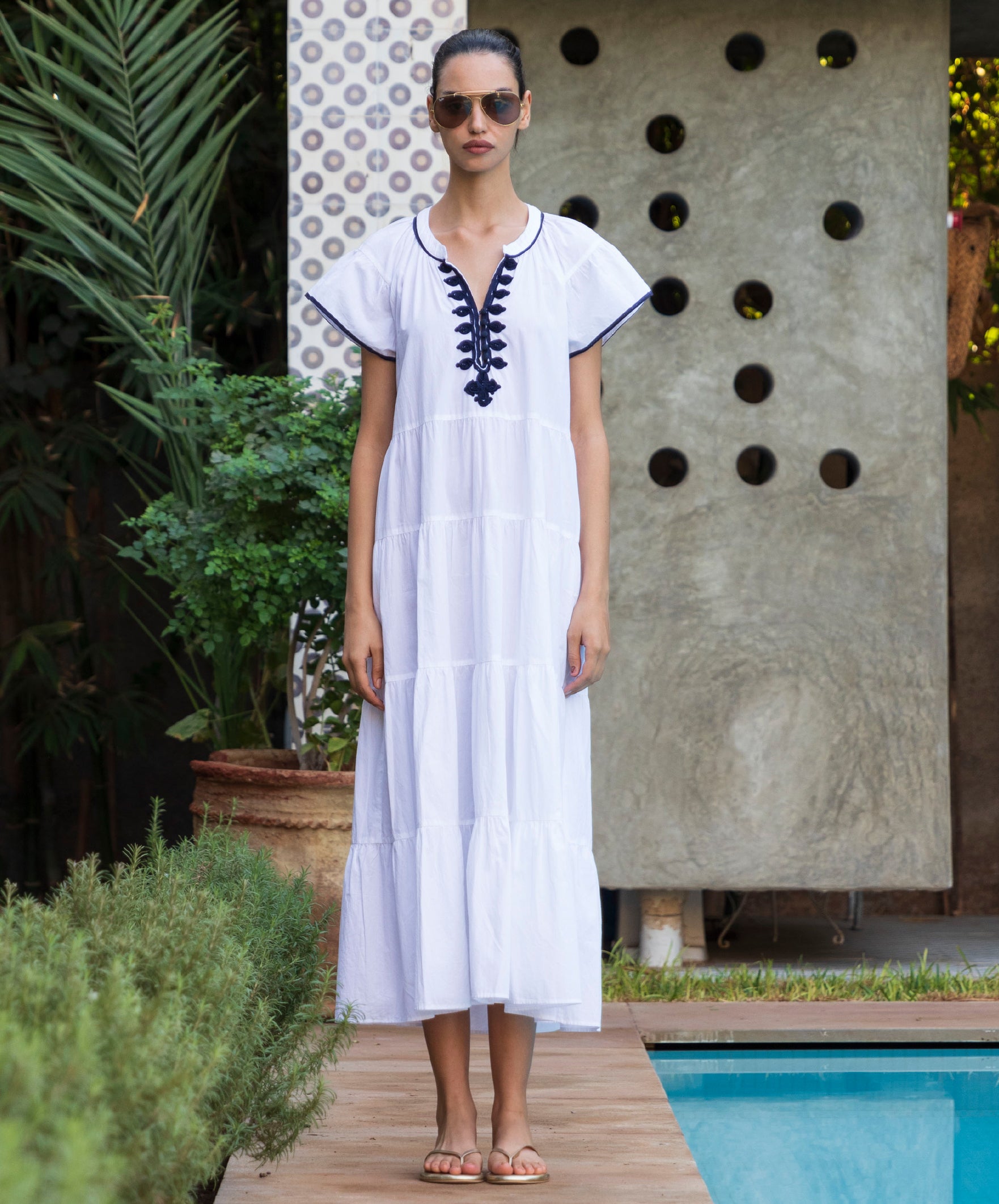 Woman in a white Camelia dress with blue passementerie embroidery standing by a poolside.