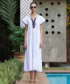 Woman in a white Camelia dress with blue passementerie embroidery standing by a poolside.