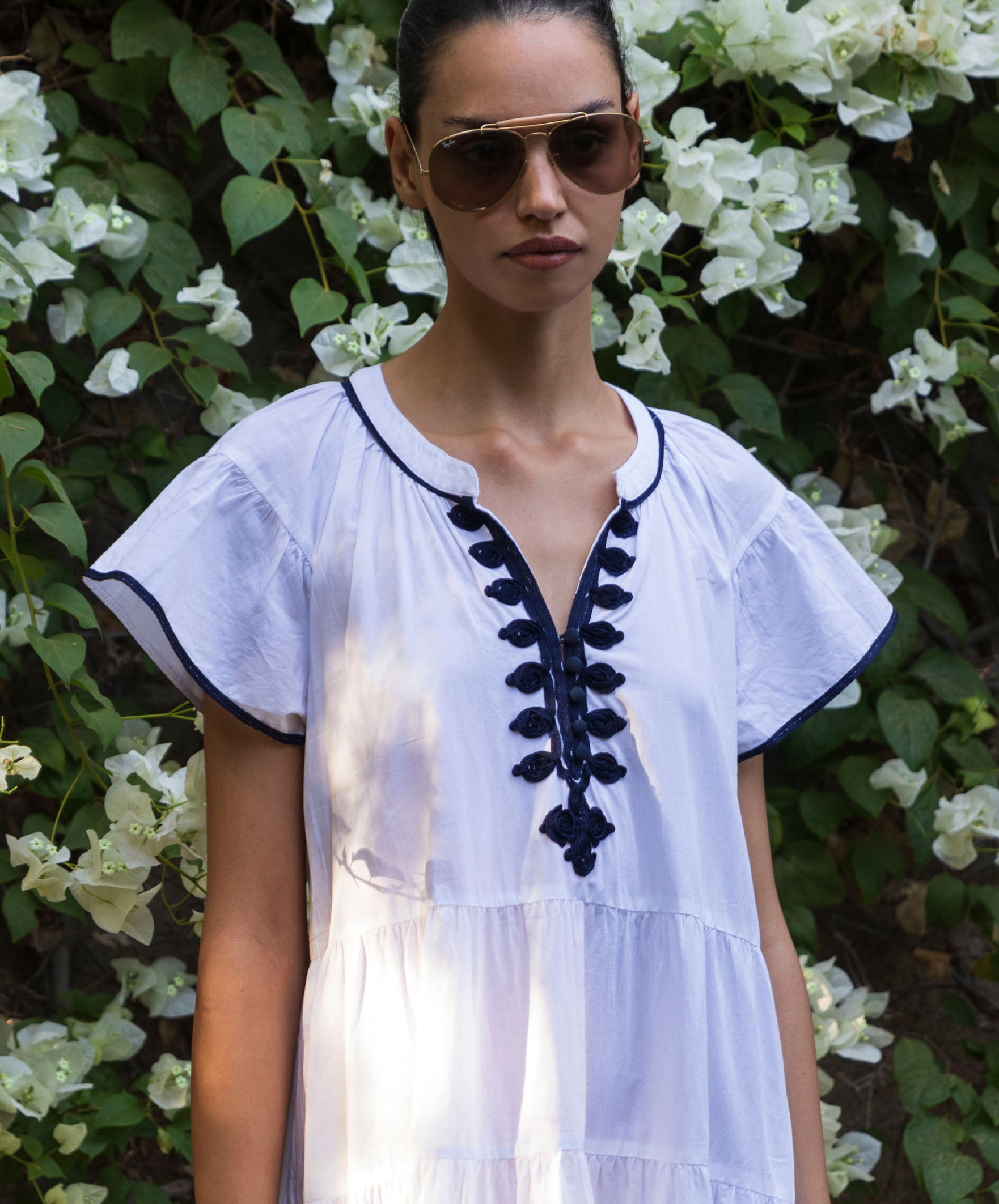 Close up of a model wearing a white Camelia dress with navy passementerie embroidery, standing against a floral background 