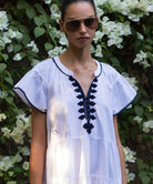 Close up of a model wearing a white Camelia dress with navy passementerie embroidery, standing against a floral background 