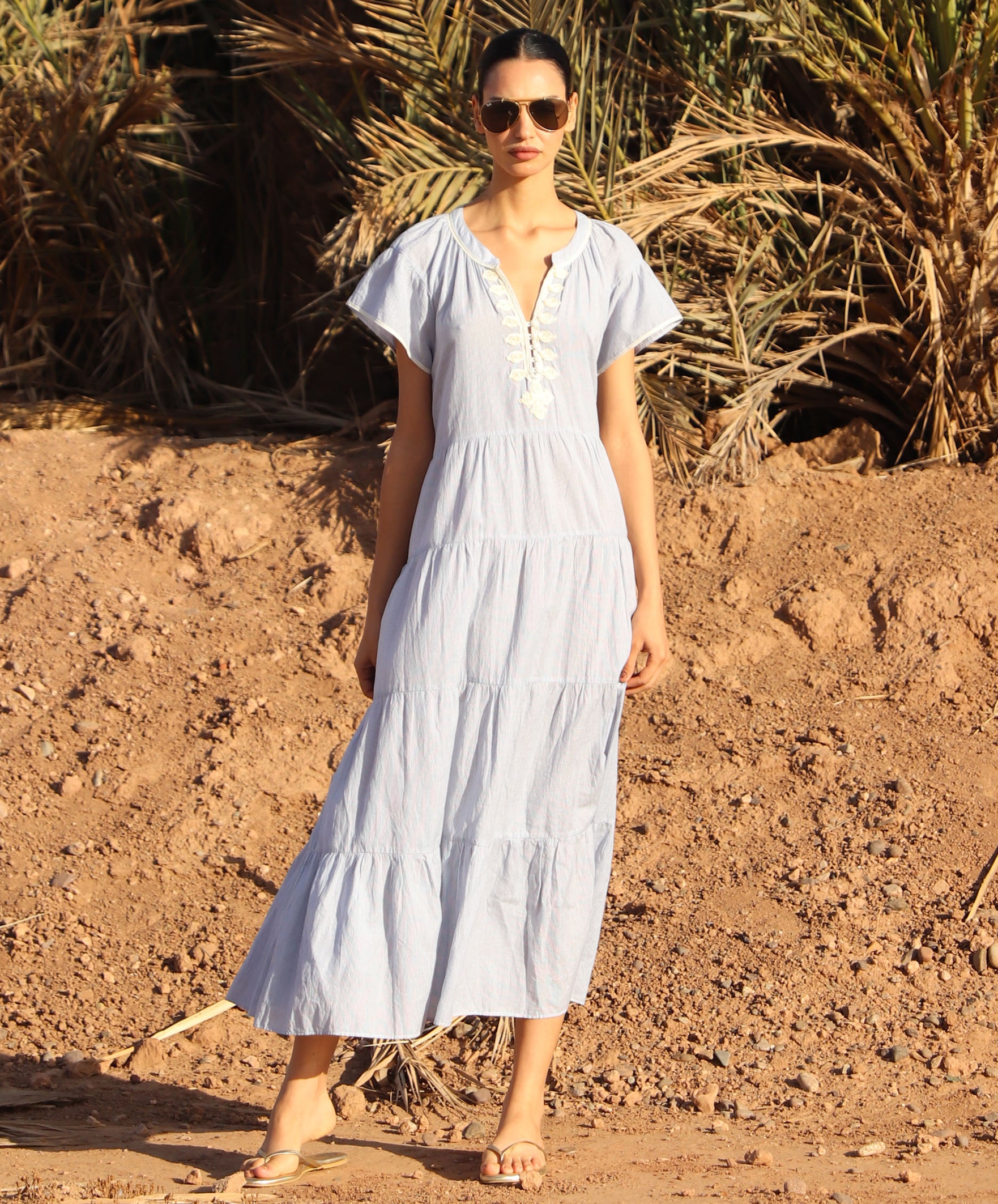 Woman in a striped blue Camelia dress standing in a desert-like setting with palm trees.