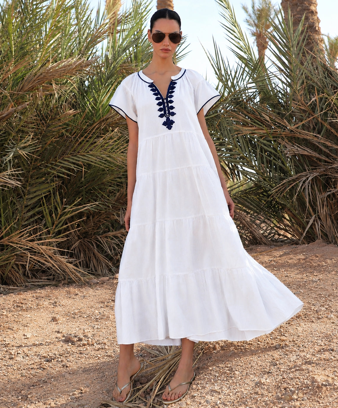 Woman in a white dress with blue embroidery standing in front of palm trees.
