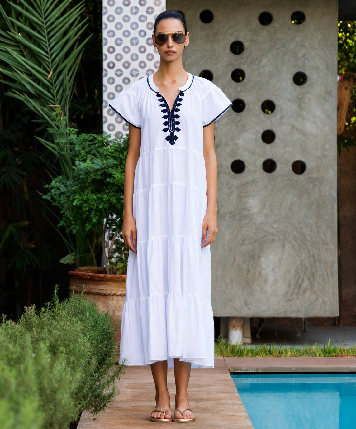 Woman in a white dress with blue embroidery standing by a poolside.