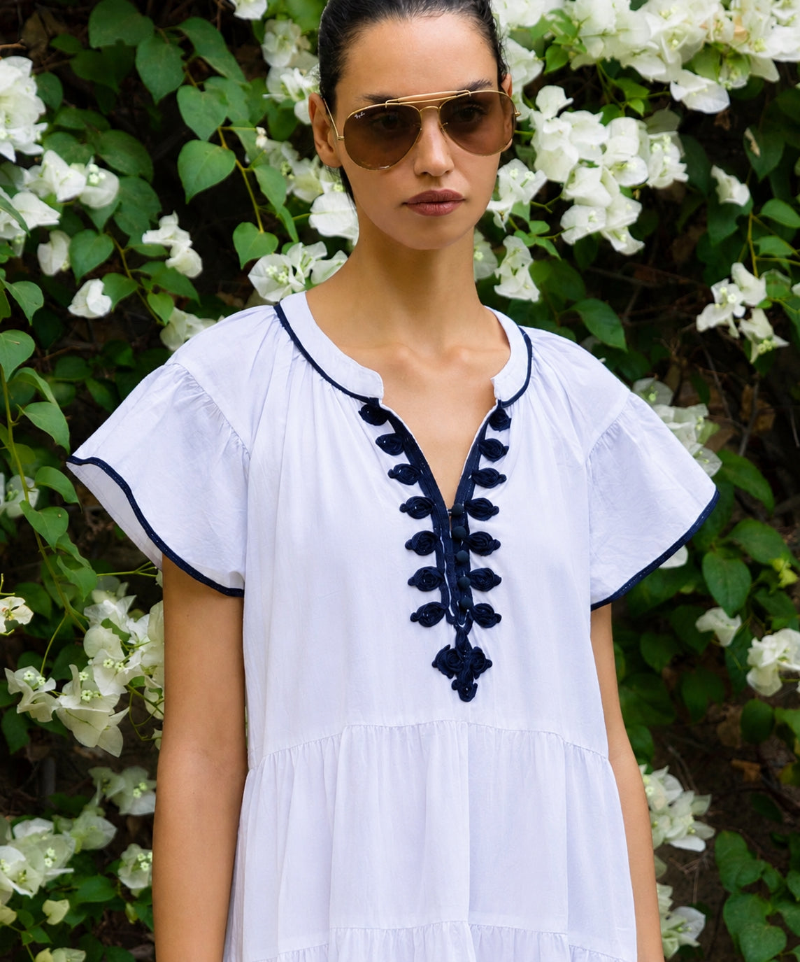 Woman wearing a white dress with black details and sunglasses, standing in front of white flowers and greenery.