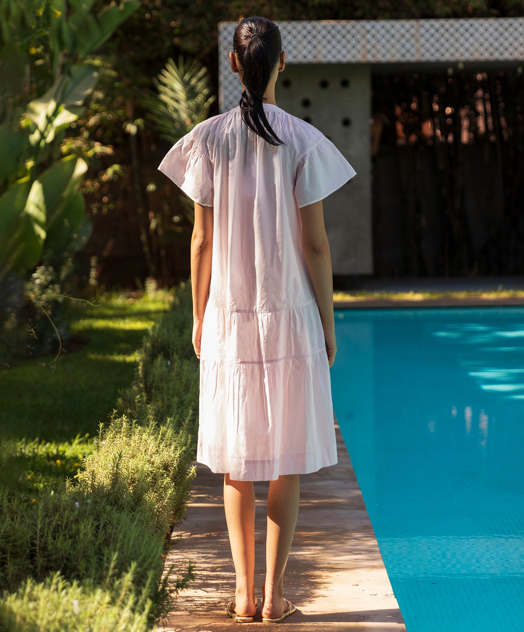 Back view of oman in a light pink Carnation dress standing by a pool with greenery around