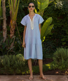 Model wearing a blue striped Carnation dress with white passementerie embroidery in a tropical garden setting