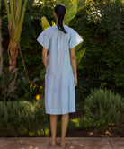 Back view of model wearing a blue striped Carnation dress standing in a tropical garden setting