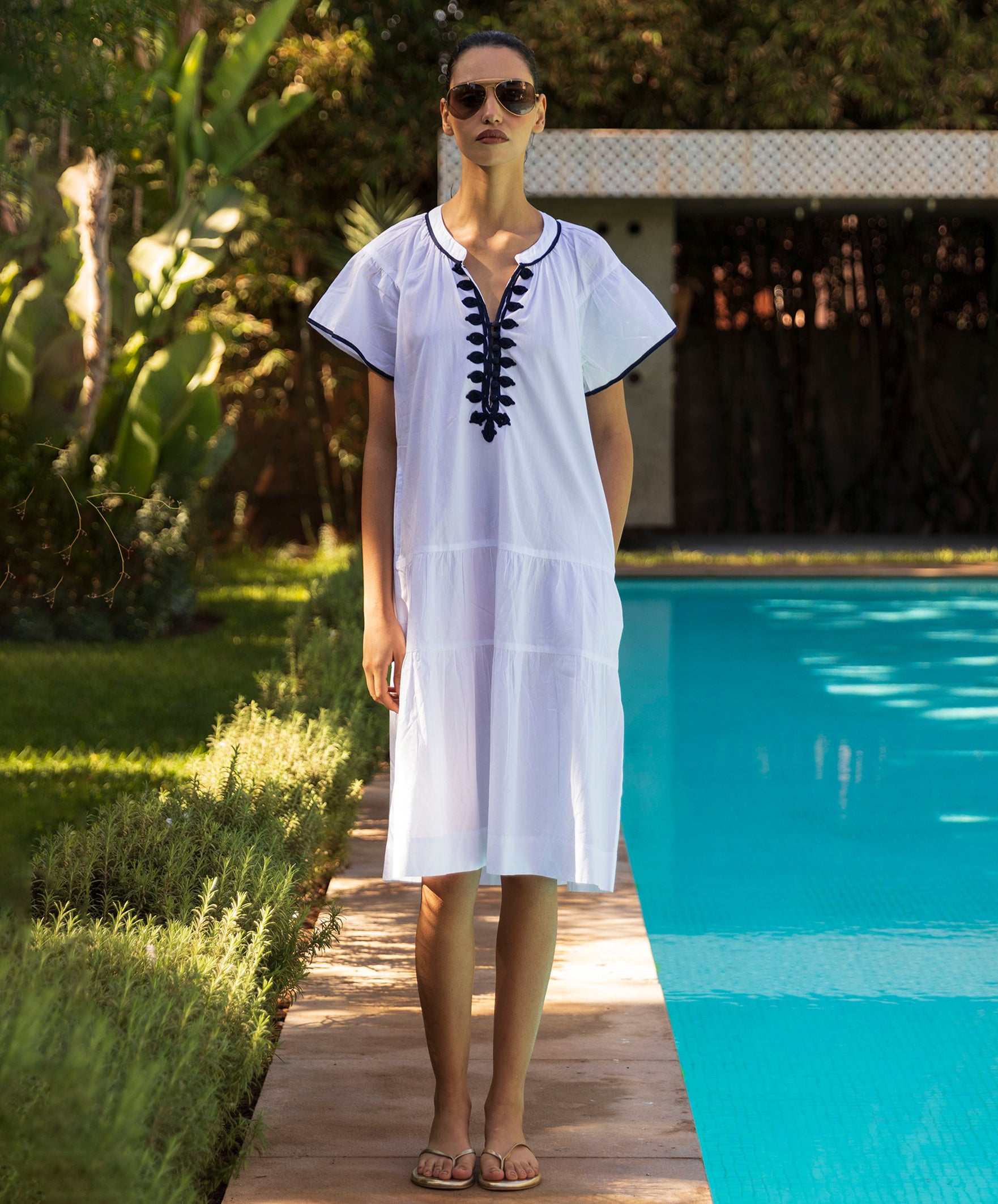 Woman in a white Carnation dress with navy passementerie embroidery standing by a pool