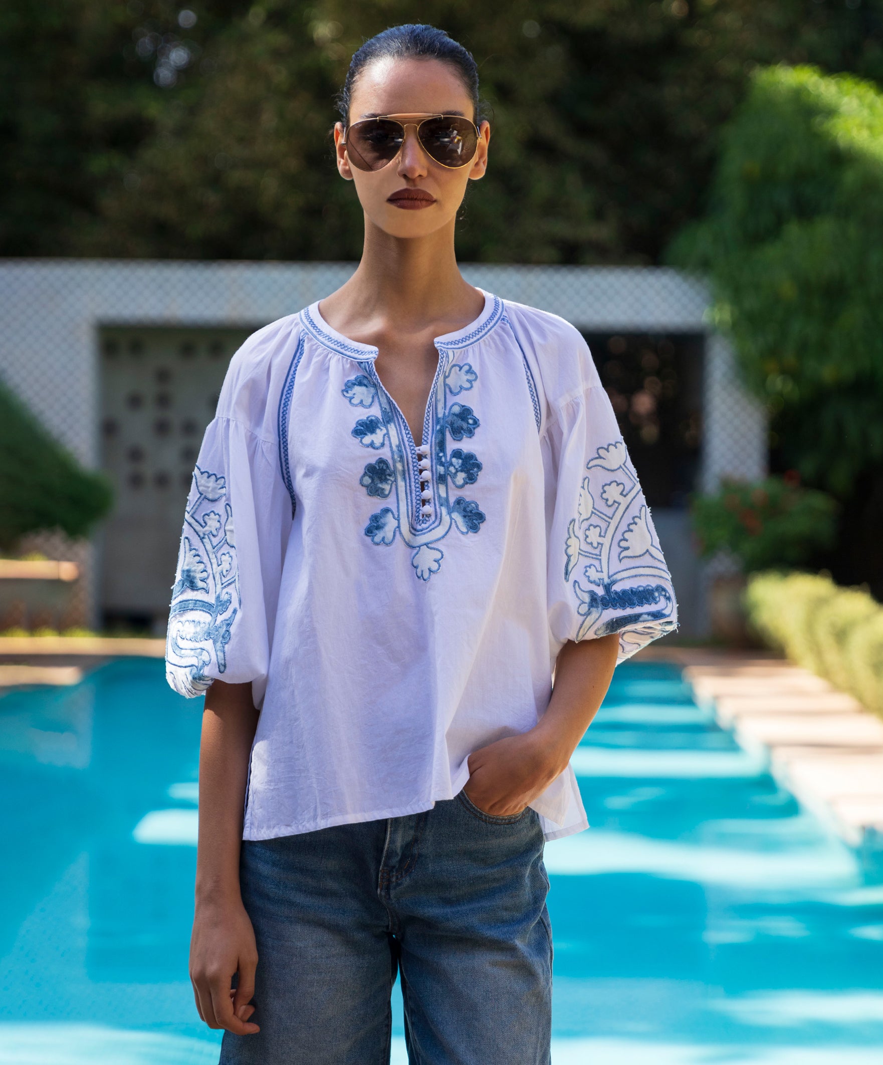 Woman wearing a white blouse with denim applique embroidered and sunglasses by a poolside.