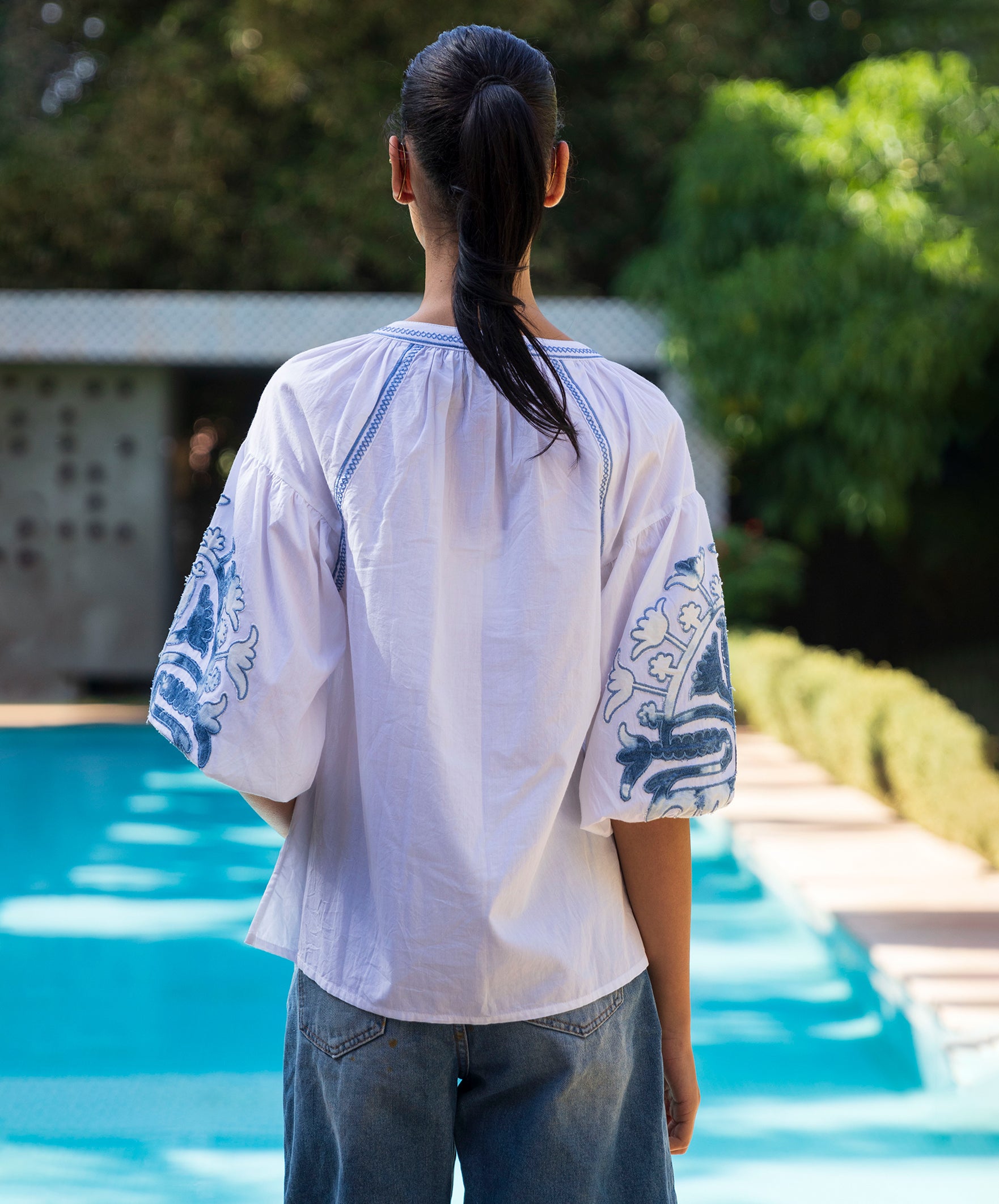 Back view of a woman wearing a white blouse with denim applique embroidery by a poolside