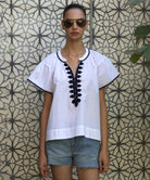 Woman wearing a white Clematis blouse with navy passementerie embroidery and denim shorts against a patterned wall.