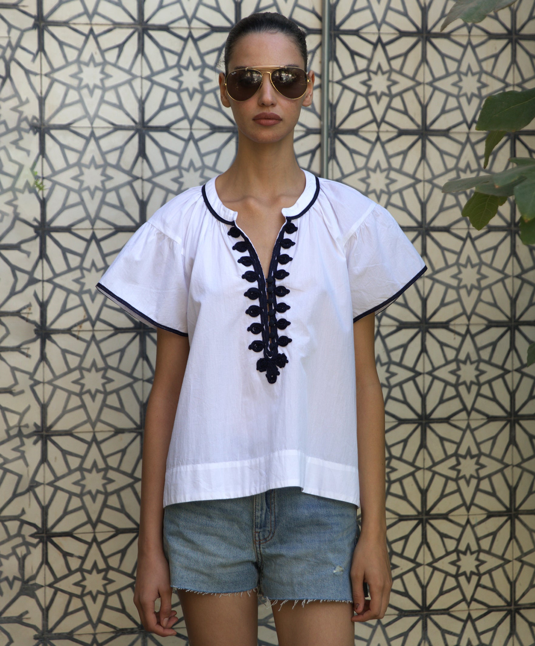 Woman wearing a white Clematis blouse with navy passementerie embroidery and denim shorts against a patterned wall.