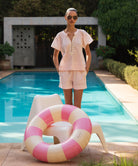 Woman in a light pink Clematis blouse and matching shorts standing by a pool with a striped pool float.