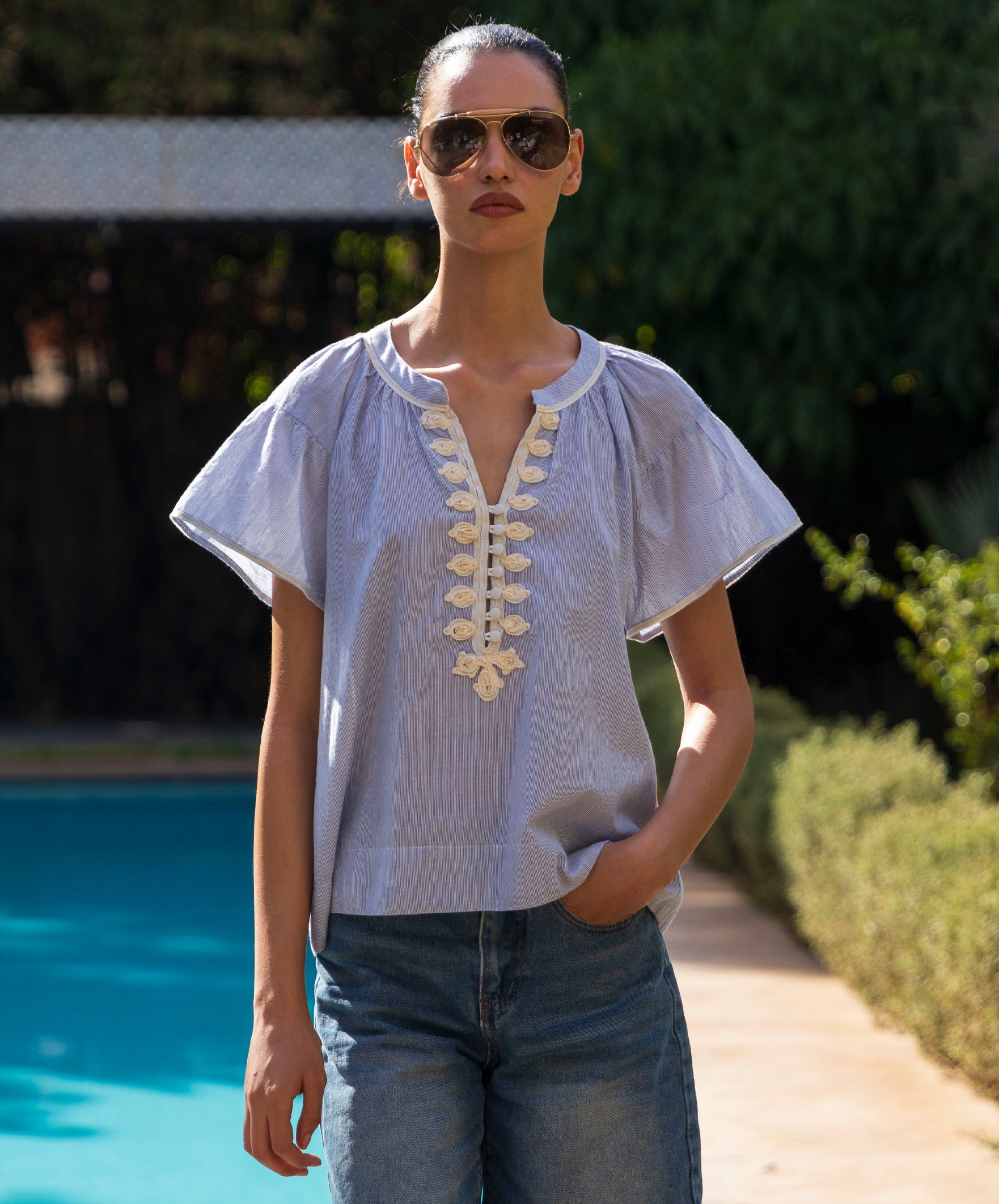 Woman wearing a blue striped blouse with passementerie embroidery details and sunglasses by a poolside.