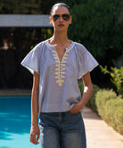 Woman wearing a blue striped blouse with passementerie embroidery details and sunglasses by a poolside.
