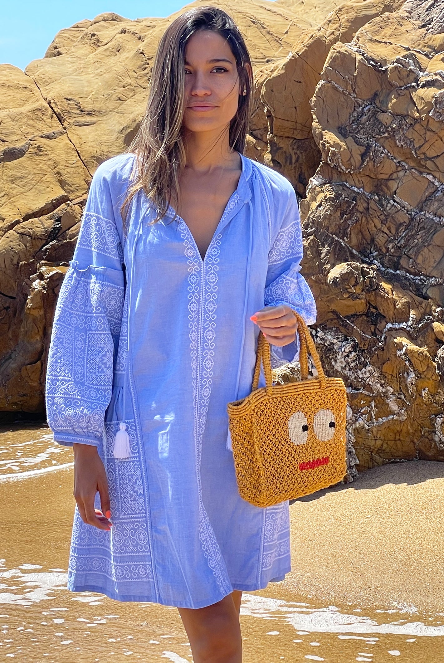 A model stood on a beach wearing a Rose and Rose blue Como dress and holding a Jacksons jute bag. 