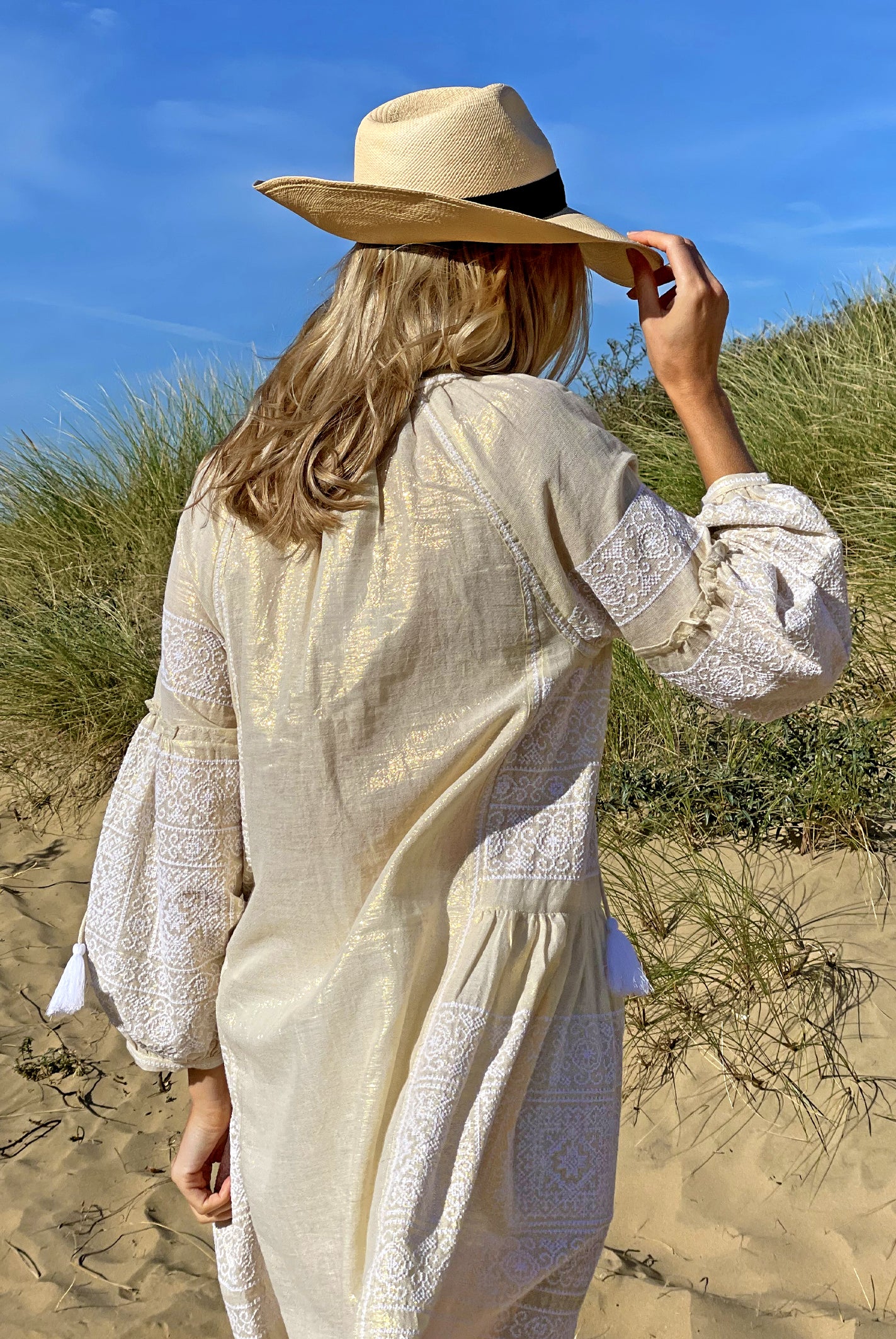 A back view of a model on a beach wearing the Rose and Rose Como dress in gold lurex and an Anthony Peto Panama hat.