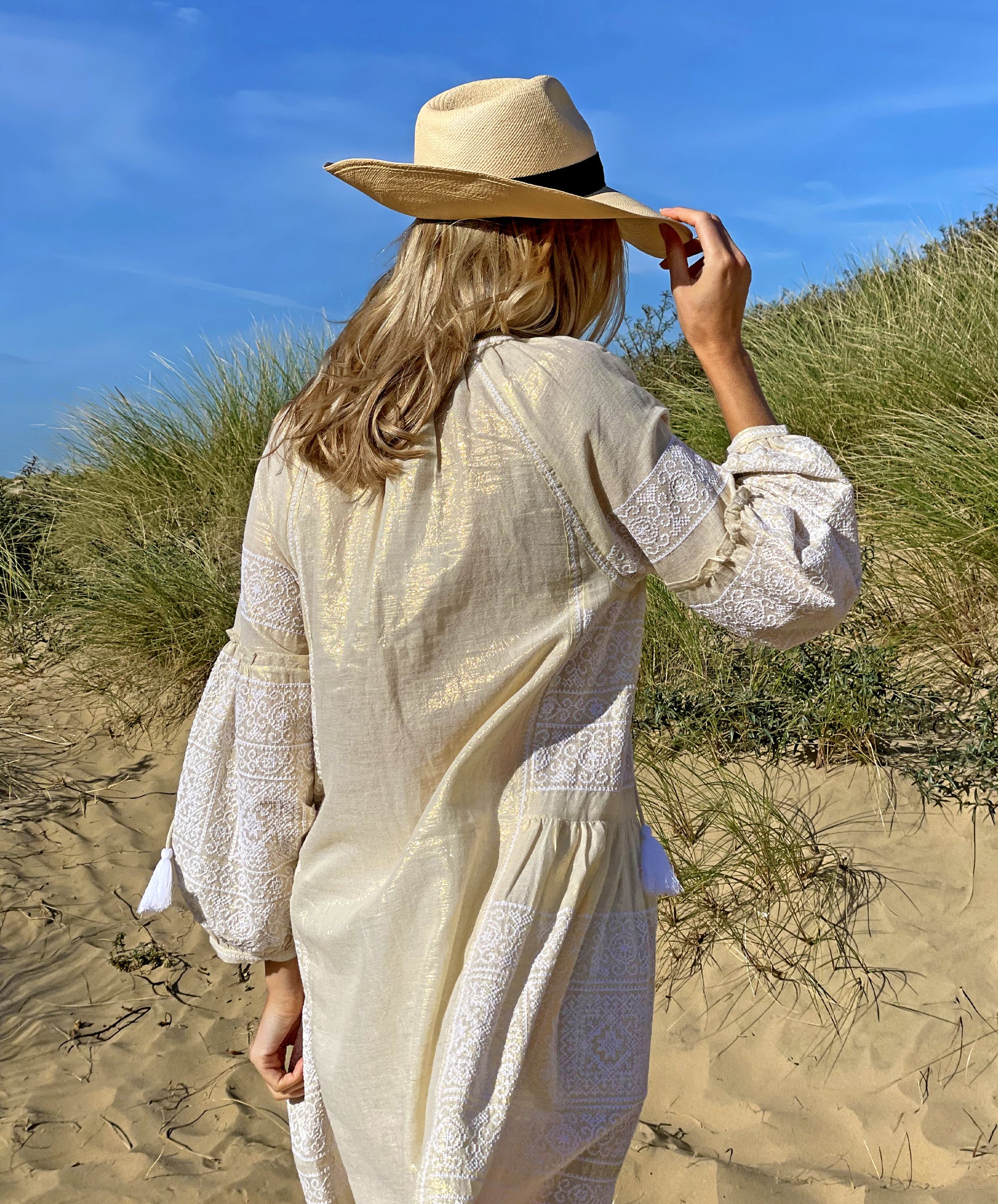 A back view of a model on a beach wearing the Rose and Rose Como dress in gold lurex and an Anthony Peto Panama hat.