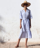 Woman in a light blue Dahlia dress and straw hat standing against a white wall