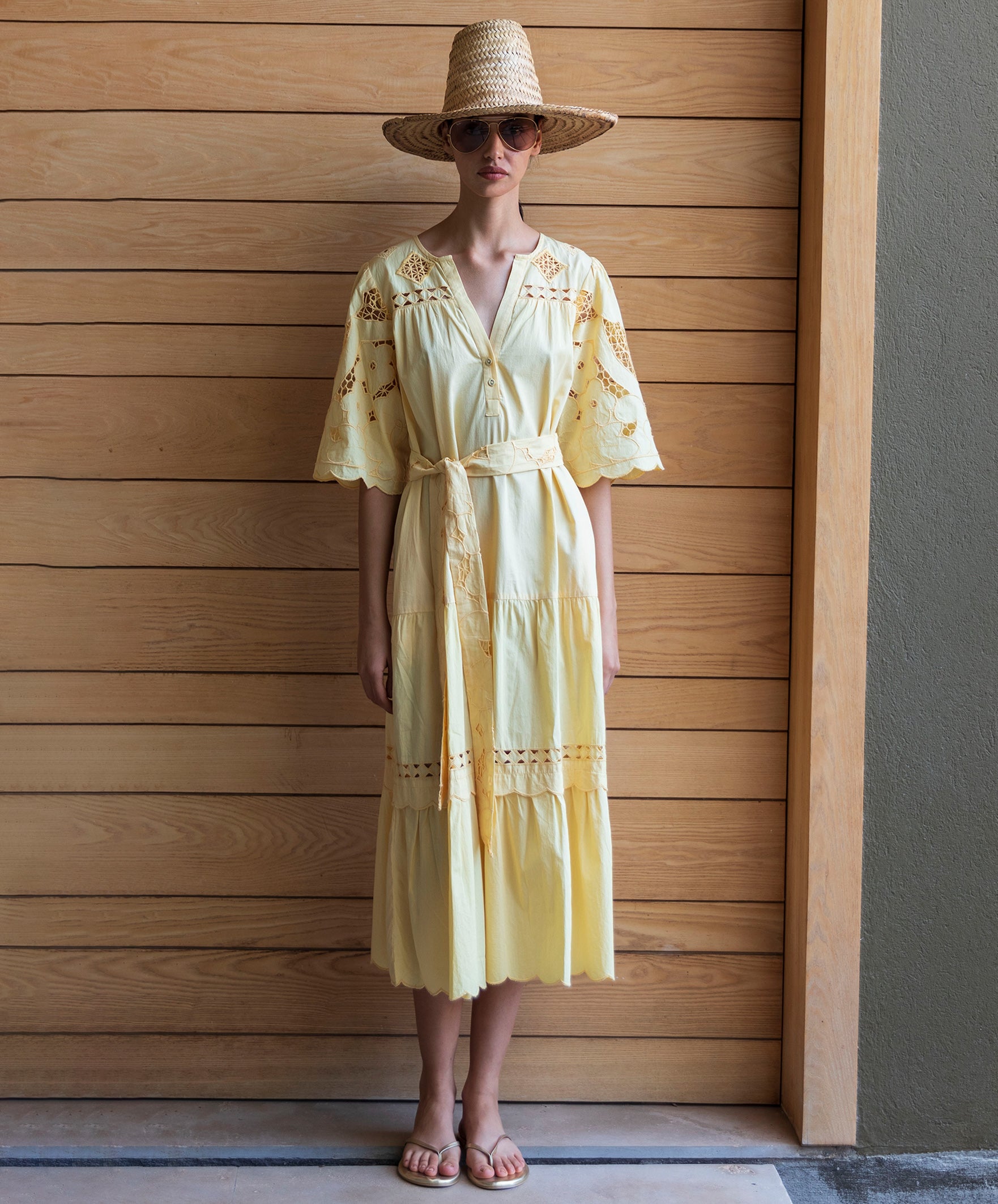 Woman wearing a light yellow embroidered dress and straw hat against a wooden wall.