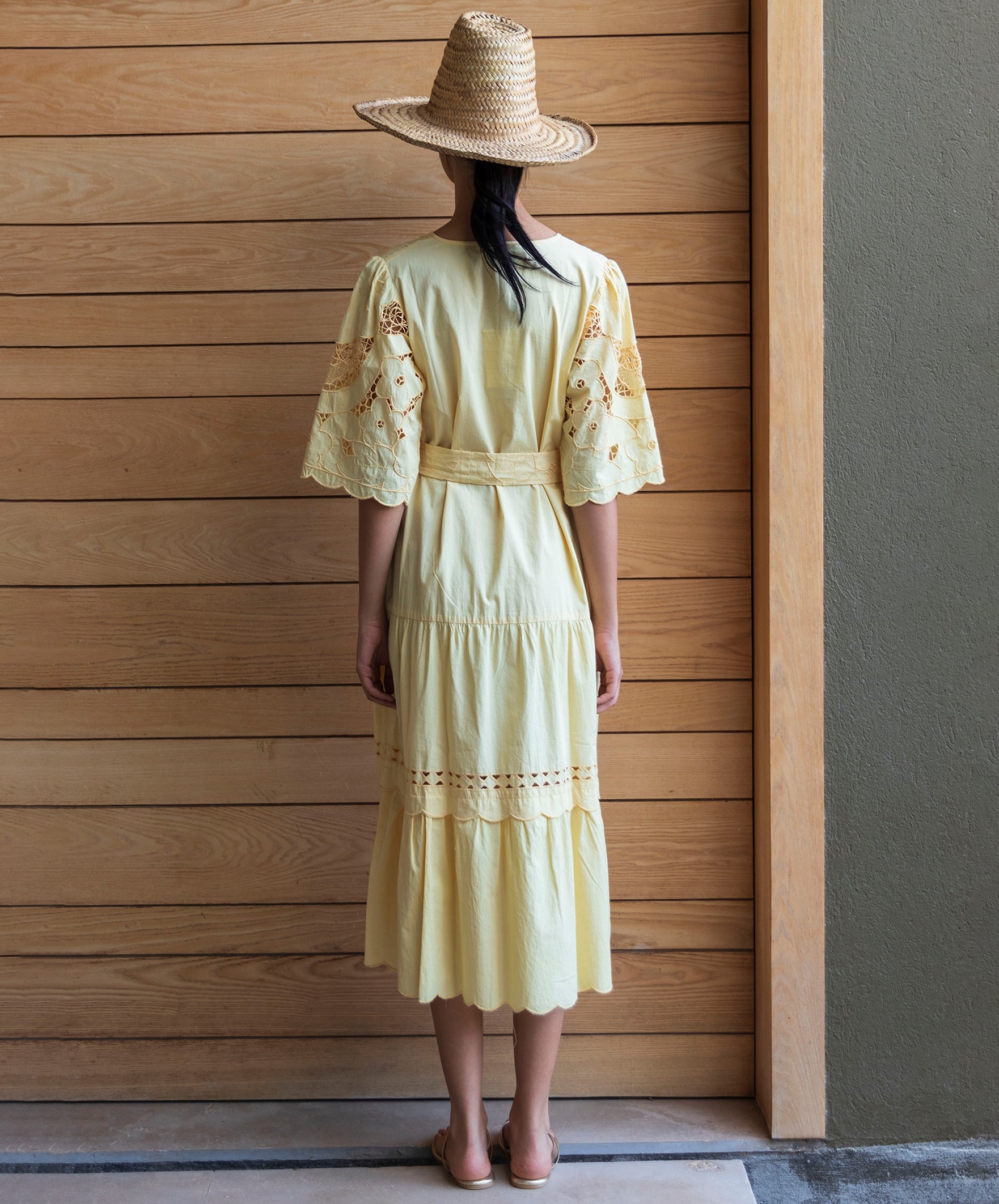 Woman wearing a light yellow embroidered dress and a straw hat, standing against a wooden wall.