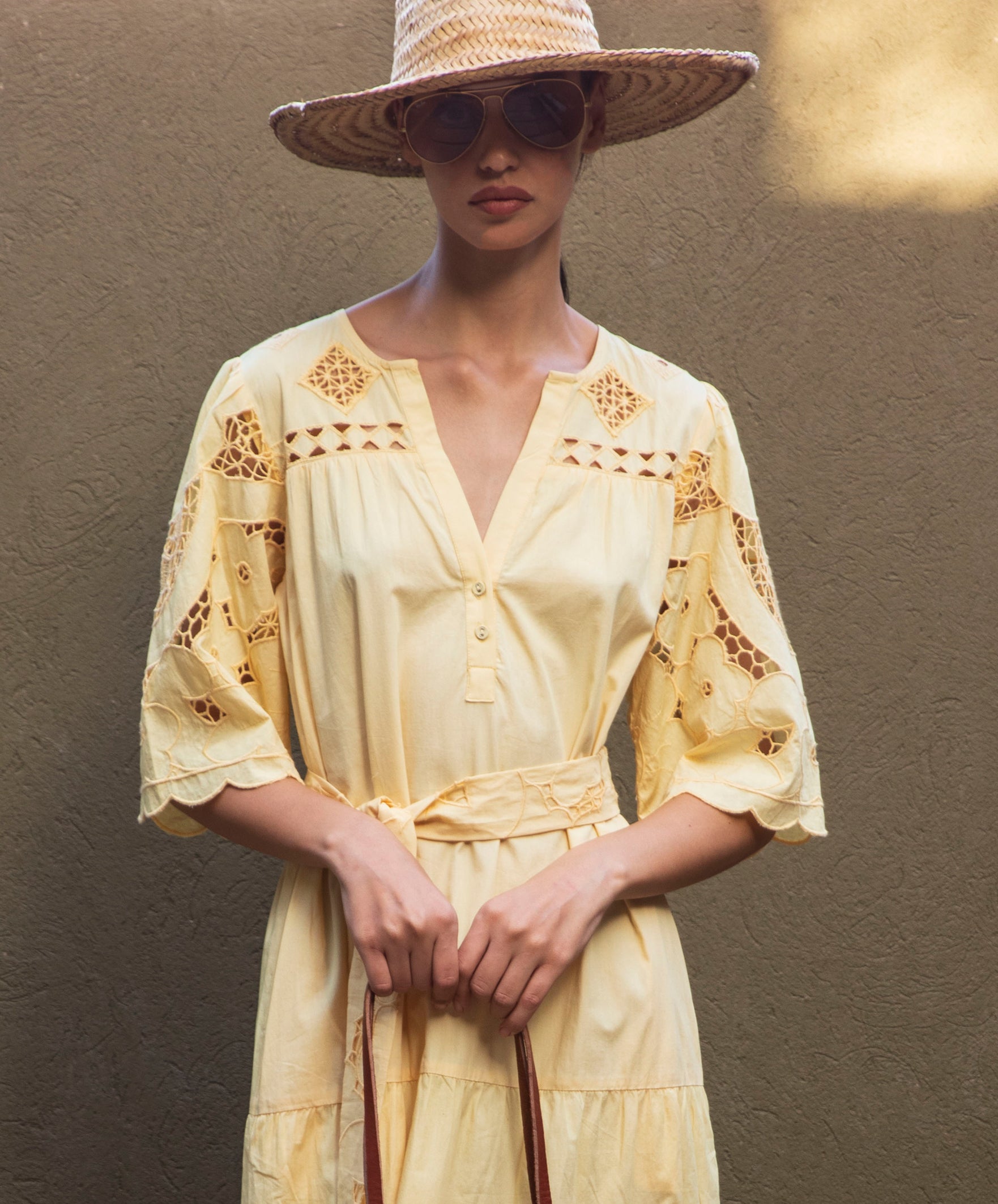 Woman wearing a pale yellow embroidered dress and a straw hat against a neutral background