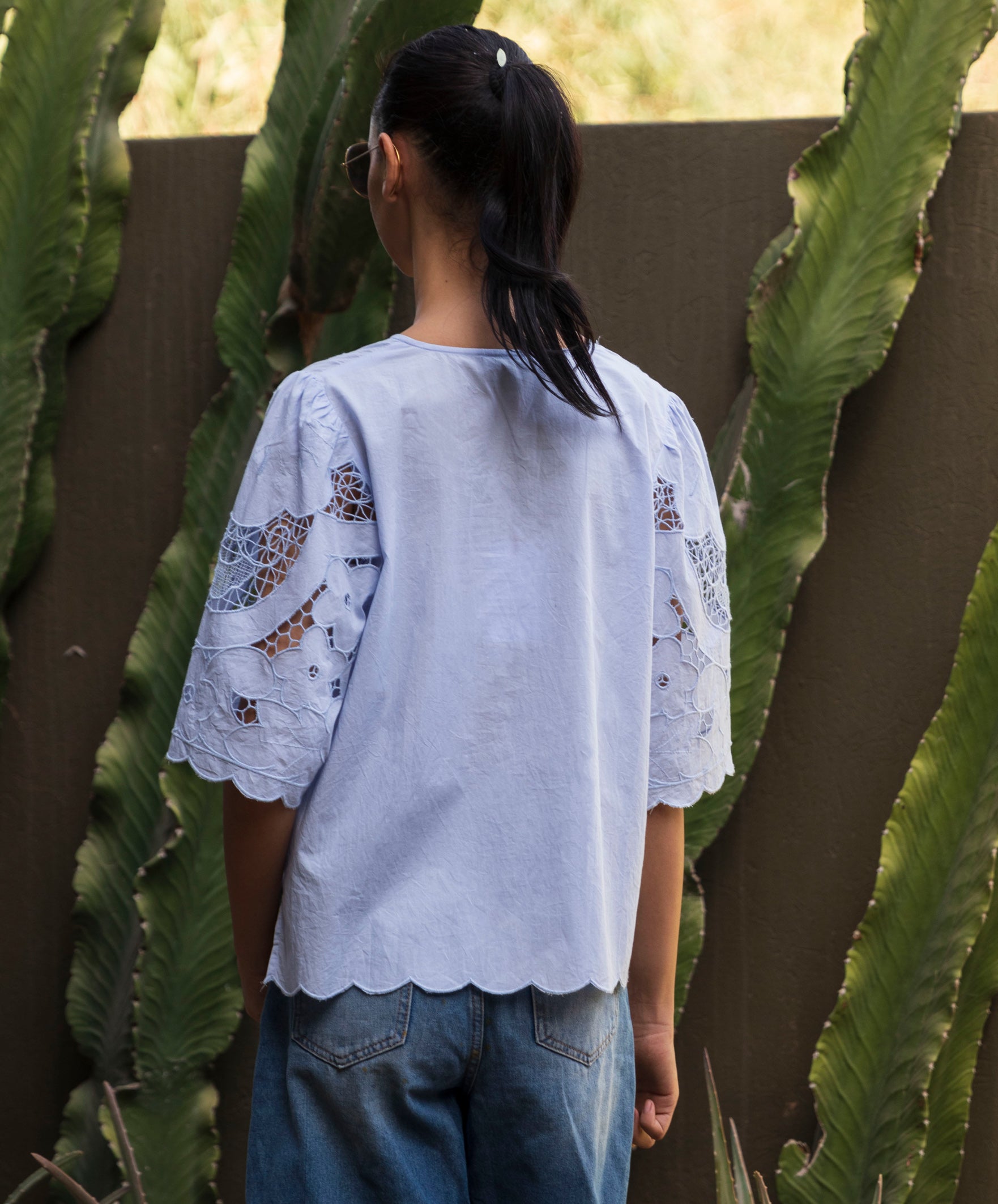 Woman wearing a light blue blouse with embroidered sleeves standing in front of cacti.