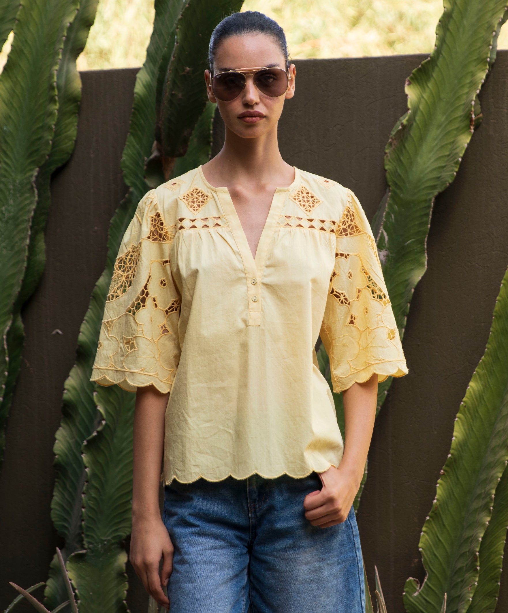 Woman wearing a pale yellow embroidered Delphinium blouse and sunglasses standing among cacti.