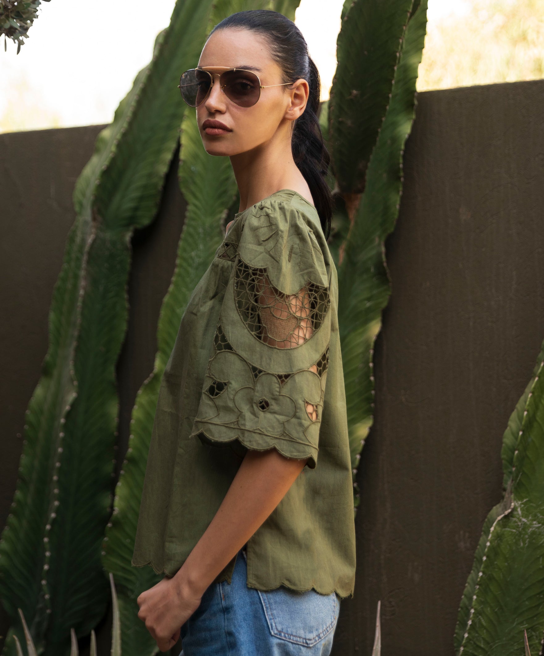 Woman wearing an olive green embroidered top and sunglasses standing in front of a cactus plant.