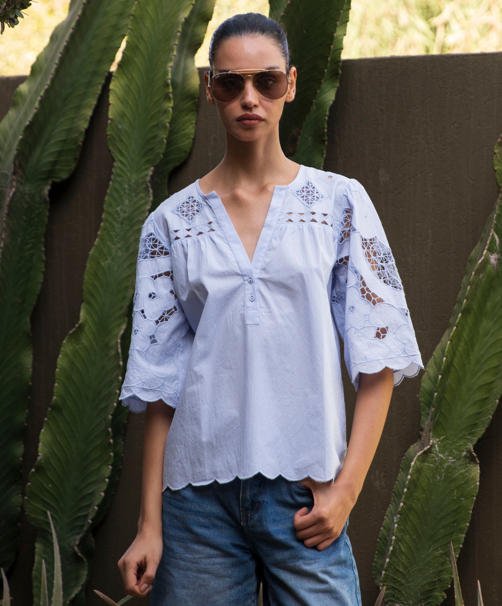 Woman wearing a light blue embroidered blouse and sunglasses standing in front of green plants.