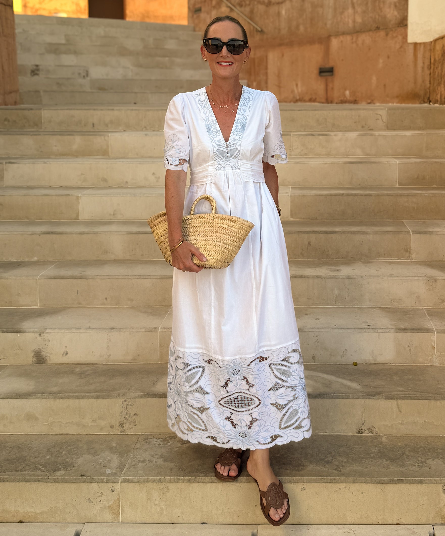 Influencer @asliceofsuffolk stands on outdoor stone steps wearing a white maxi dress with blue embroidery, holding a woven straw tote bag.