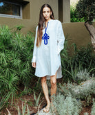 A model stood in a garden wearing a Rose and Rose Euston white linen tunic shirt with cobalt blue passementerie decoration.