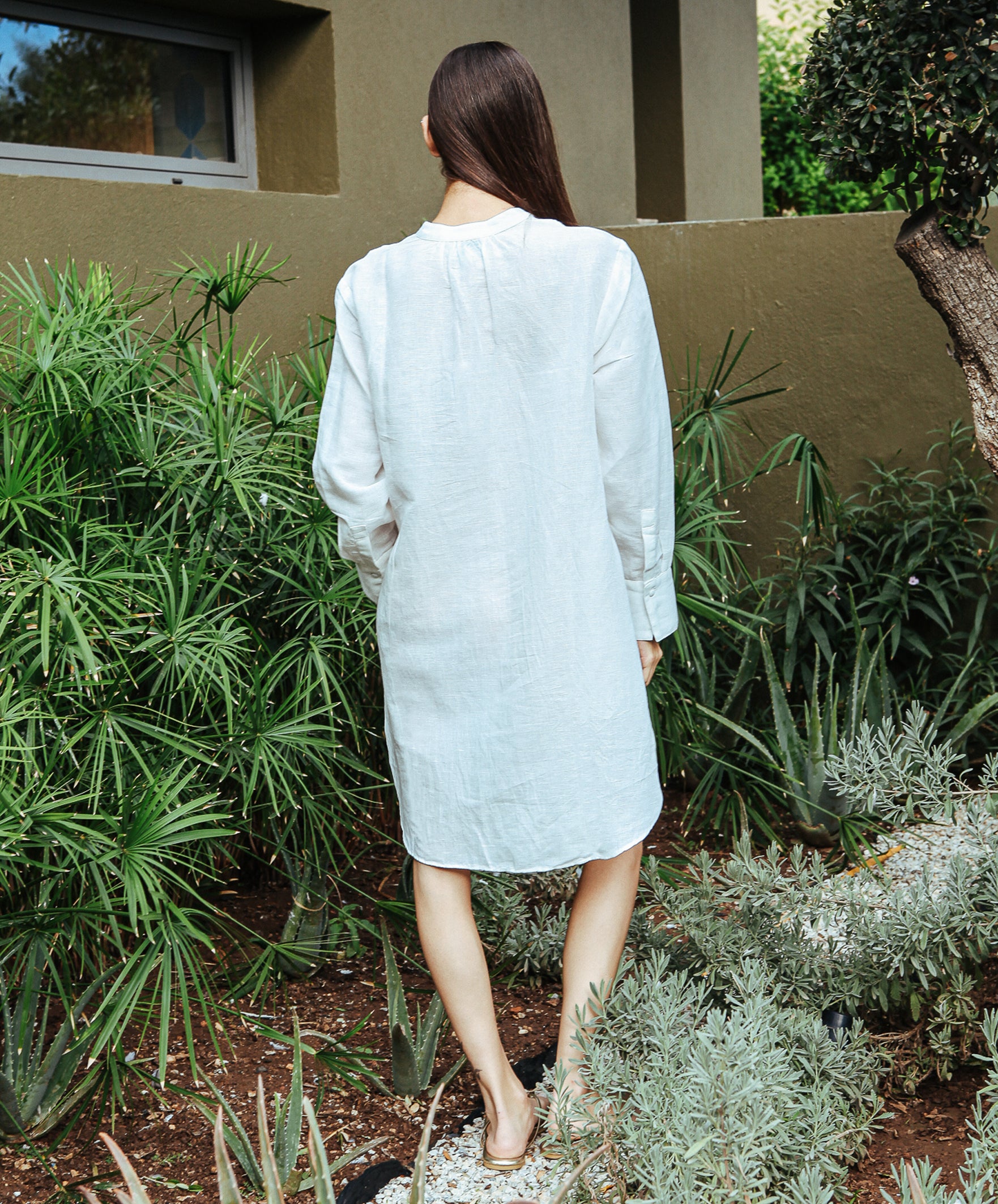 A back view of a model stood in a garden wearing a Rose and Rose Euston white linen tunic shirt.