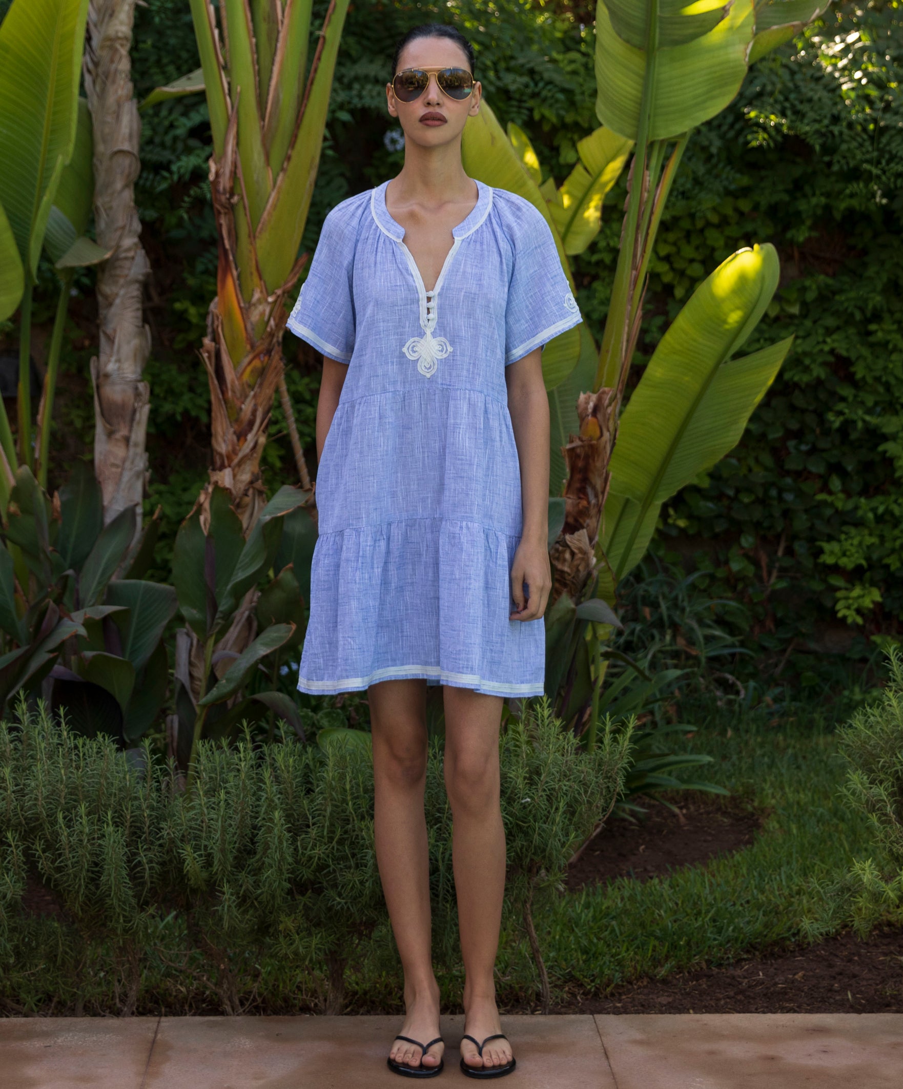 Woman wearing a light blue Flora dress with white passementerie decoration standing in a tropical garden setting