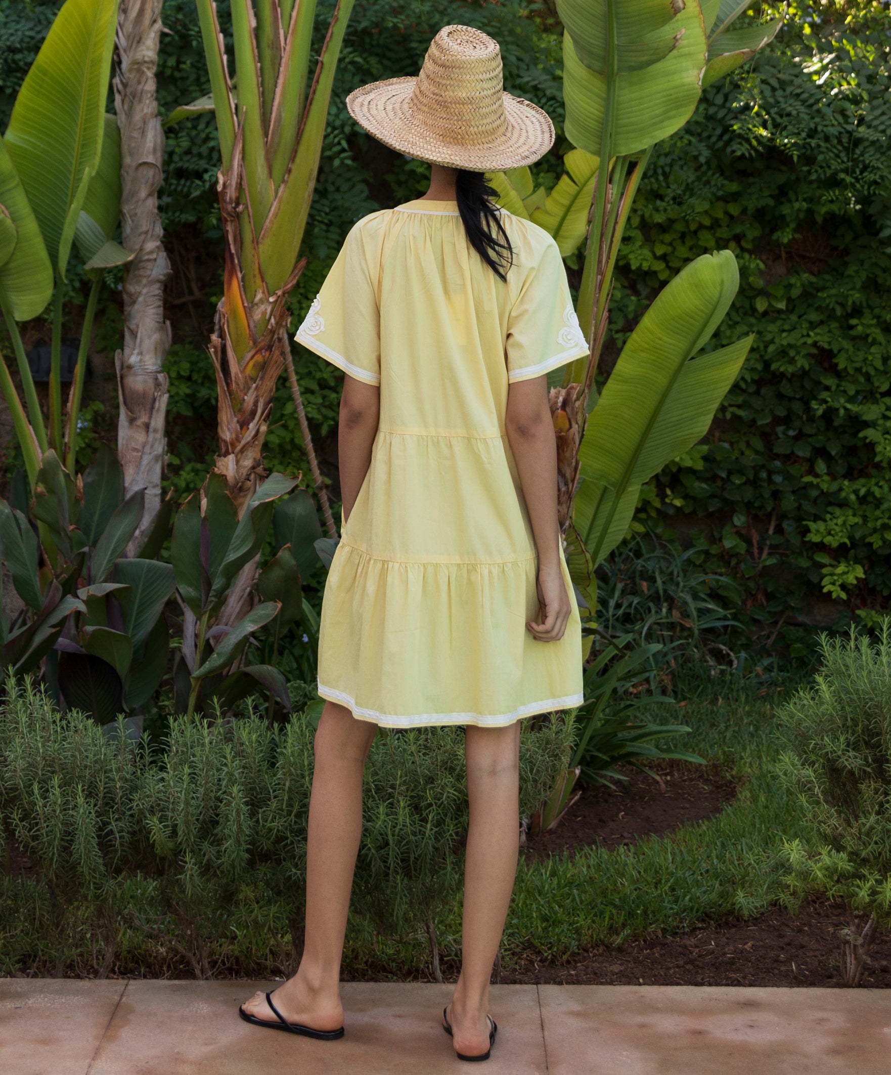 Back view of a woman wearing a pale yellow Flora dress and straw hat standing in a tropical garden setting