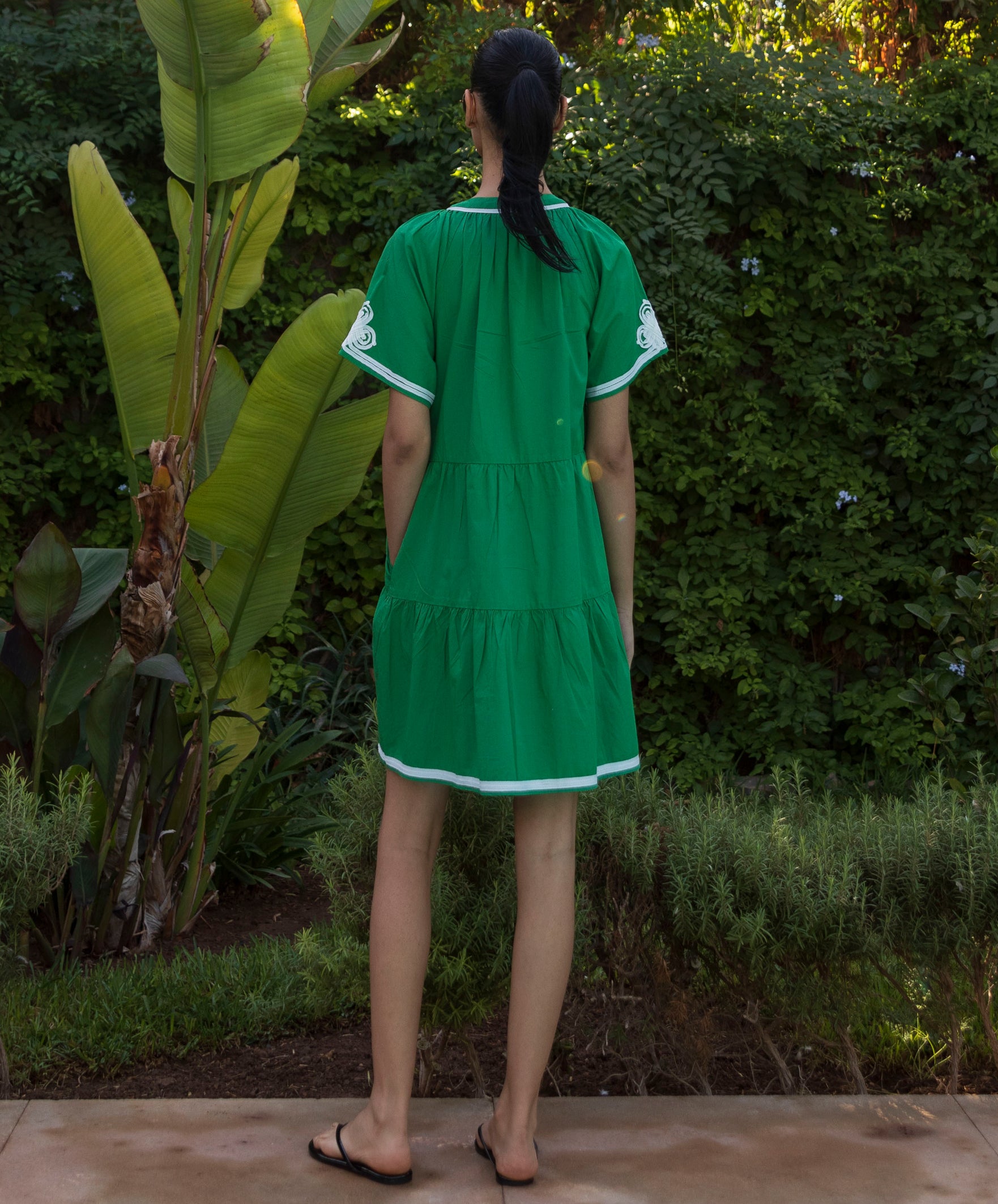 Back view of woman wearing a green Flora dress with white passementerie trim standing in a tropical garden setting.
