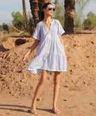 Woman in a light blue Flora dress standing in a desert-like setting with palm trees.