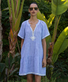 Woman wearing a light blue Flora dress with white passementerie trim standing among tropical plants