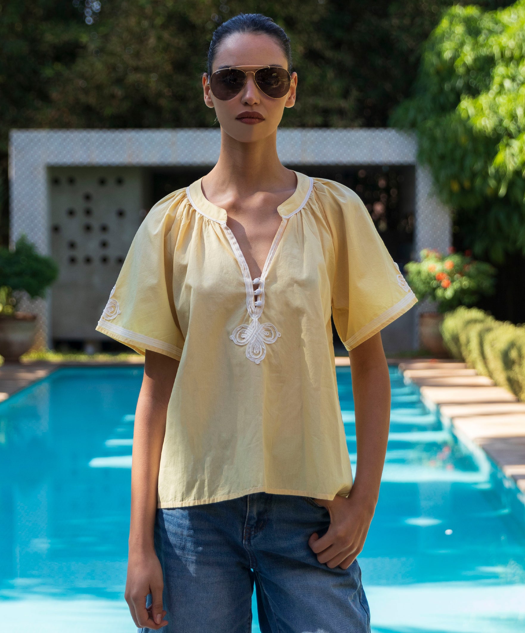 Woman wearing a pale yellow Foxglove blouse with passementerie decoration and sunglasses by a pool.