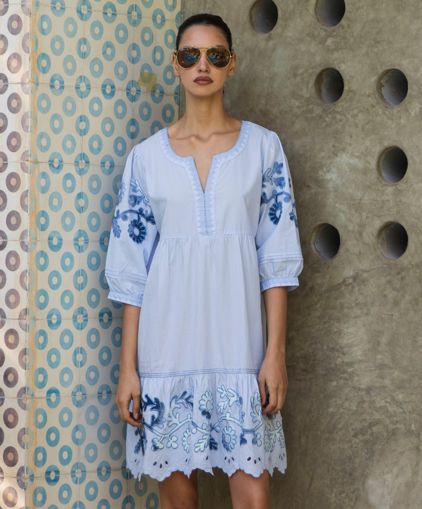 Woman wearing a light blue dress with floral embroidery against a textured wall.