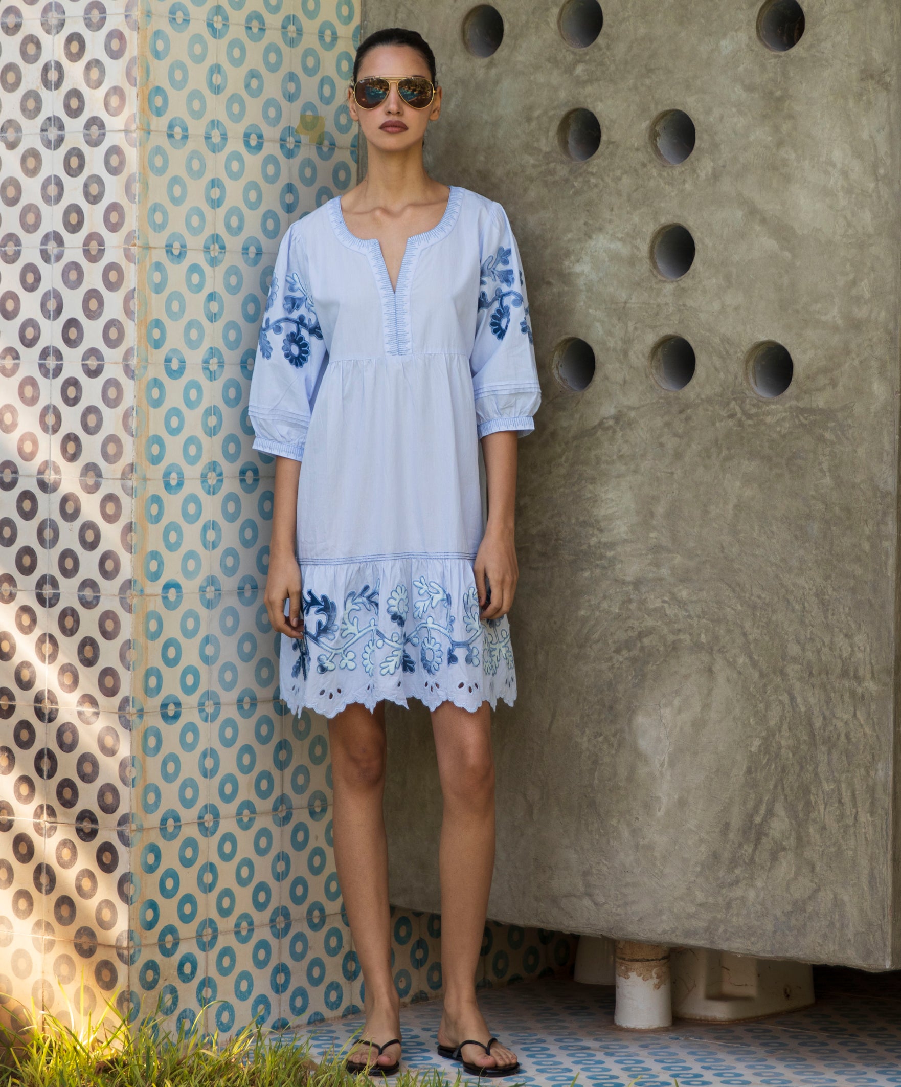 Woman wearing a light blue dress with floral embroidery standing against a textured wall.