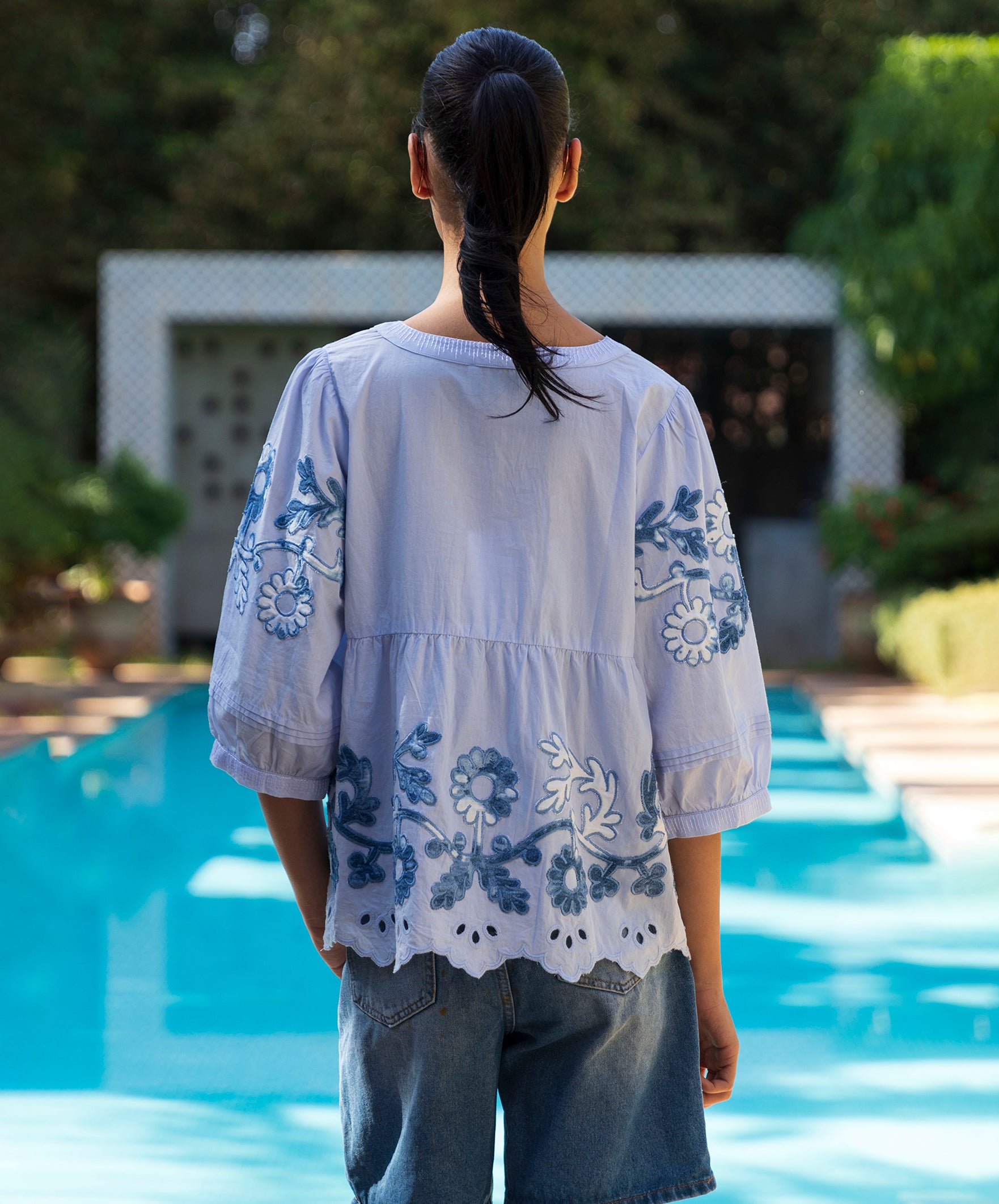 Woman wearing a light blue embroidered top in front of a pool