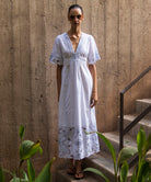 Woman in a white maxi dress with blue embroidery standing on steps with a textured wall background