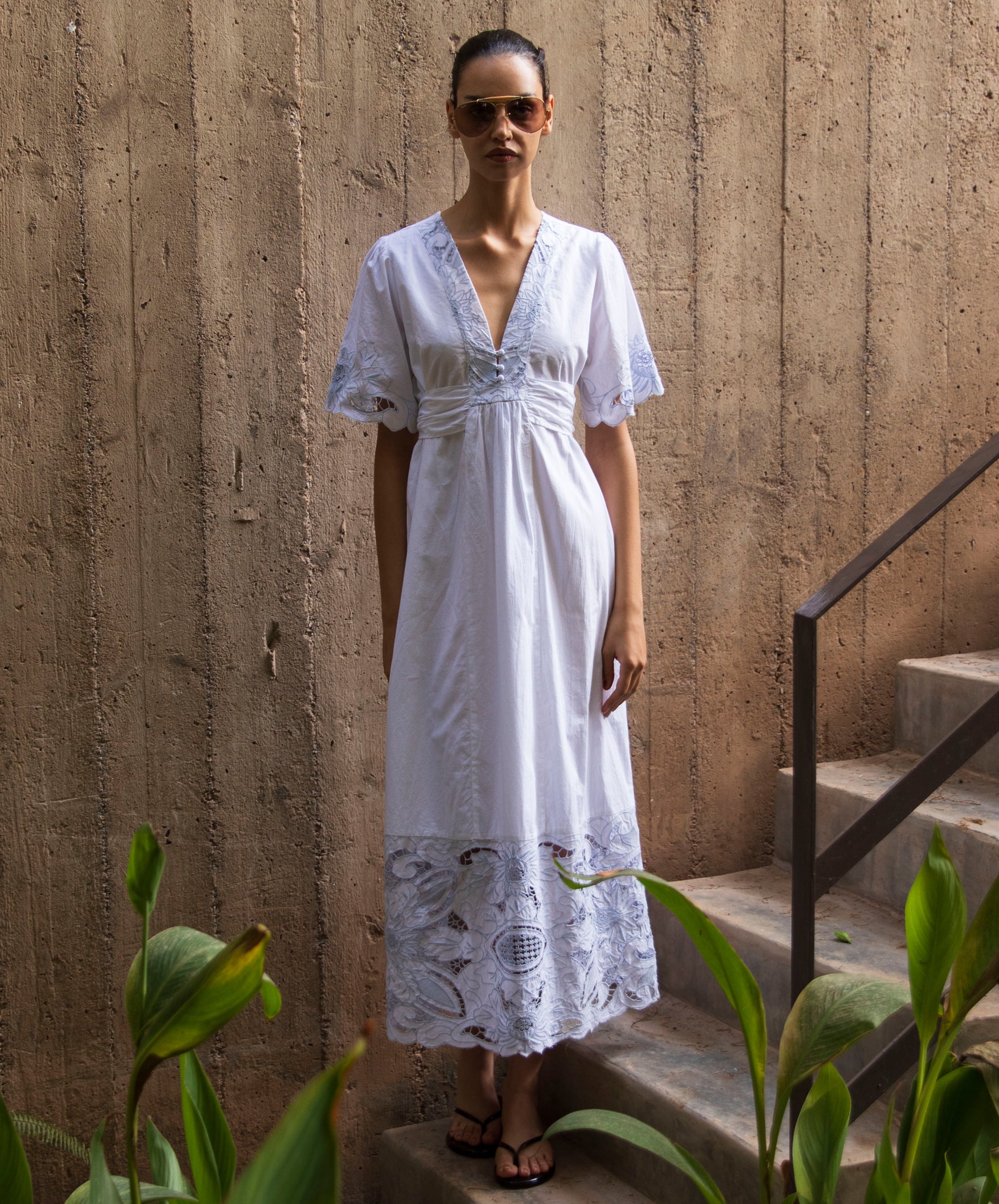 Woman in a white maxi dress with blue embroidery standing on steps with a textured wall background